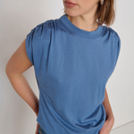 A woman wearing a slate blue short-sleeve top with gathered puff sleeves and blue jeans, photographed against a plain off-white background.