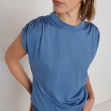 A woman wearing a slate blue short-sleeve top with gathered puff sleeves and blue jeans, photographed against a plain off-white background.