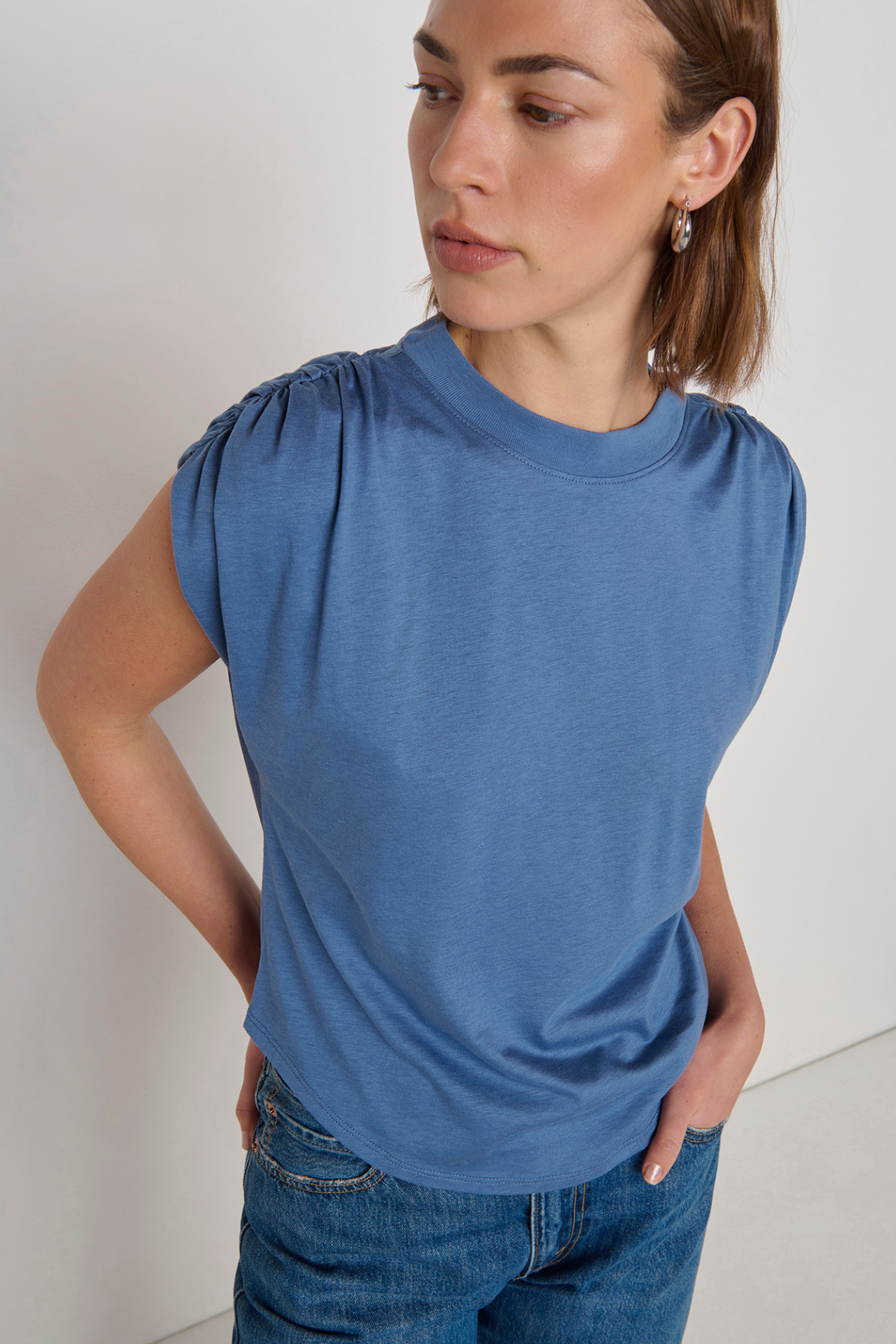 A woman wearing a slate blue short-sleeve top with gathered puff sleeves and blue jeans, photographed against a plain off-white background.