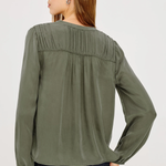 A woman shown from the back wearing an olive green long-sleeve blouse with pleated detailing across the upper back, paired with dark blue jeans, against a plain light gray studio background.