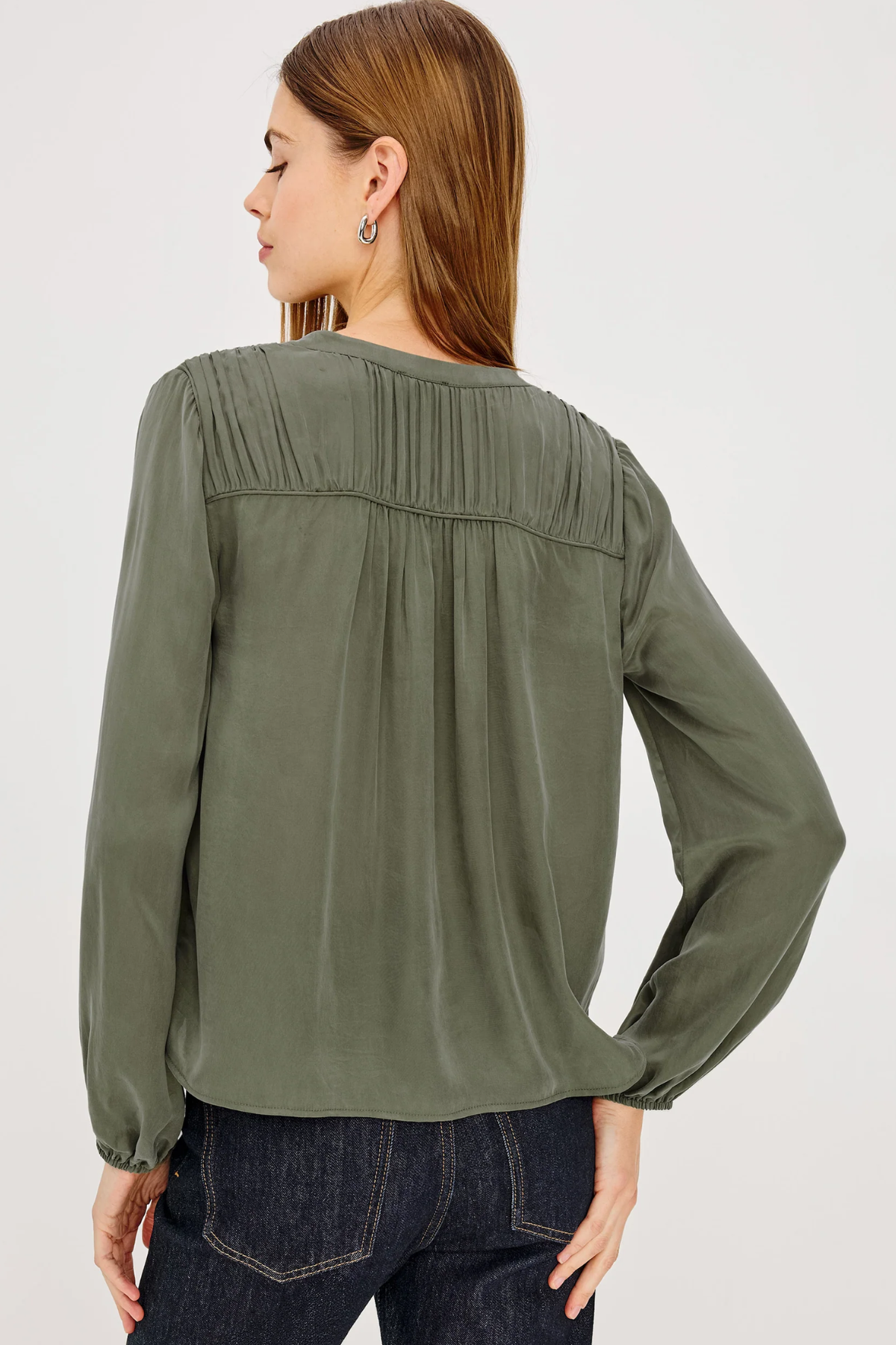 A woman shown from the back wearing an olive green long-sleeve blouse with pleated detailing across the upper back, paired with dark blue jeans, against a plain light gray studio background.