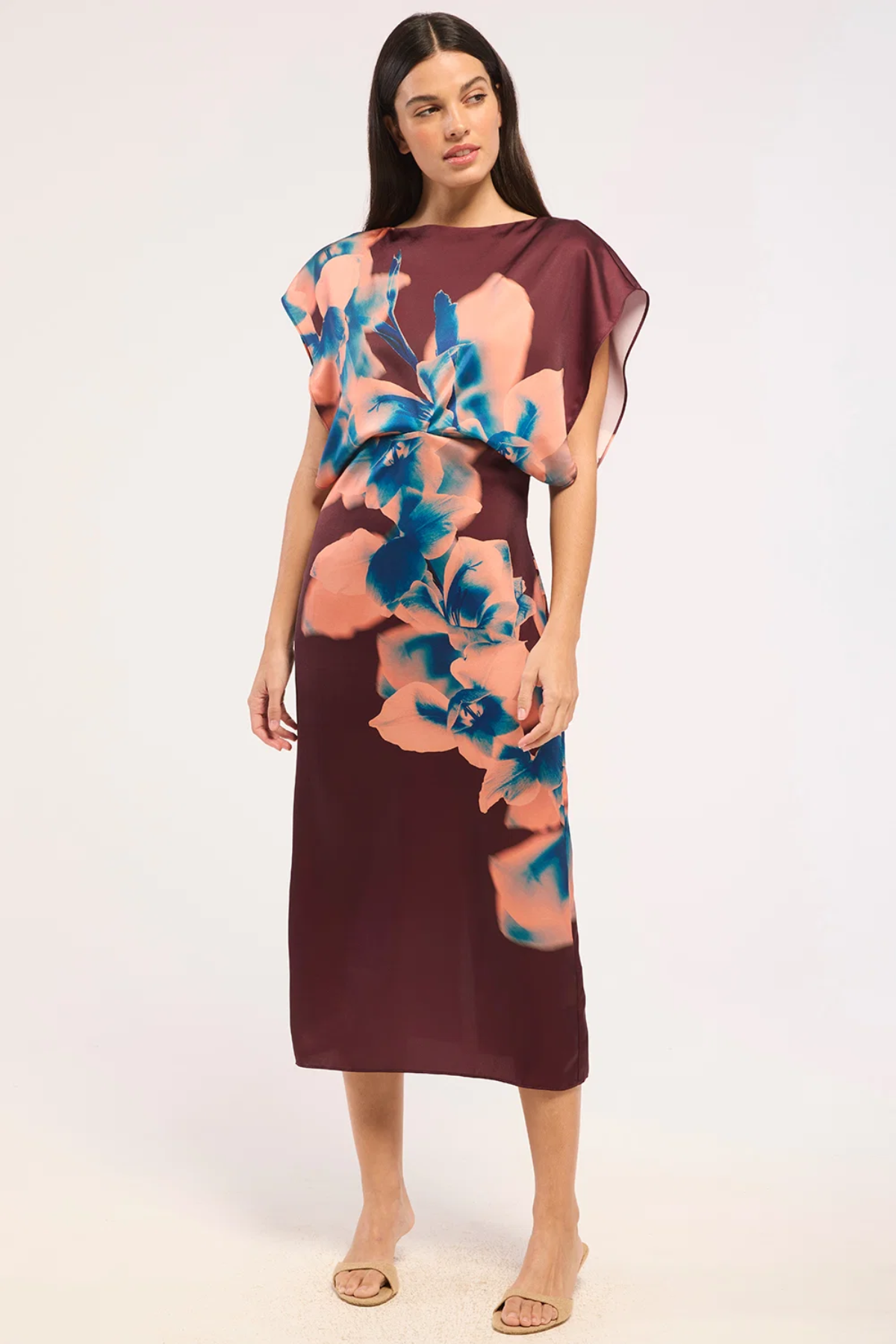 A woman modeling a sleeveless midi dress with a deep V-neckline, draped sheer cap sleeves, and a large floral print in peach and blue over a dark burgundy base, set against a plain white studio background.