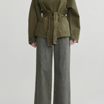 A model wearing an olive green belted jacket with a high mock neck and wide-leg gray jeans, standing against a light gray studio background.