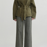 A model wearing an olive green belted jacket with a high mock neck and wide-leg gray jeans, standing against a light gray studio background.