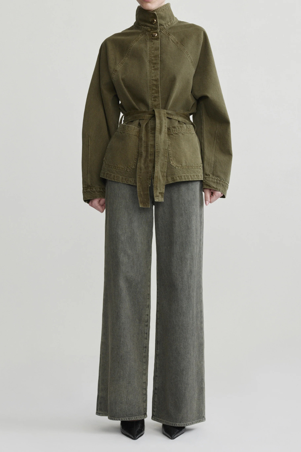 A model wearing an olive green belted jacket with a high mock neck and wide-leg gray jeans, standing against a light gray studio background.