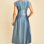 A woman shown from the back wearing a light blue midi dress with cap sleeves, a gathered waist, and a satin-like finish, paired with tan high-heeled sandals against a plain beige studio background.