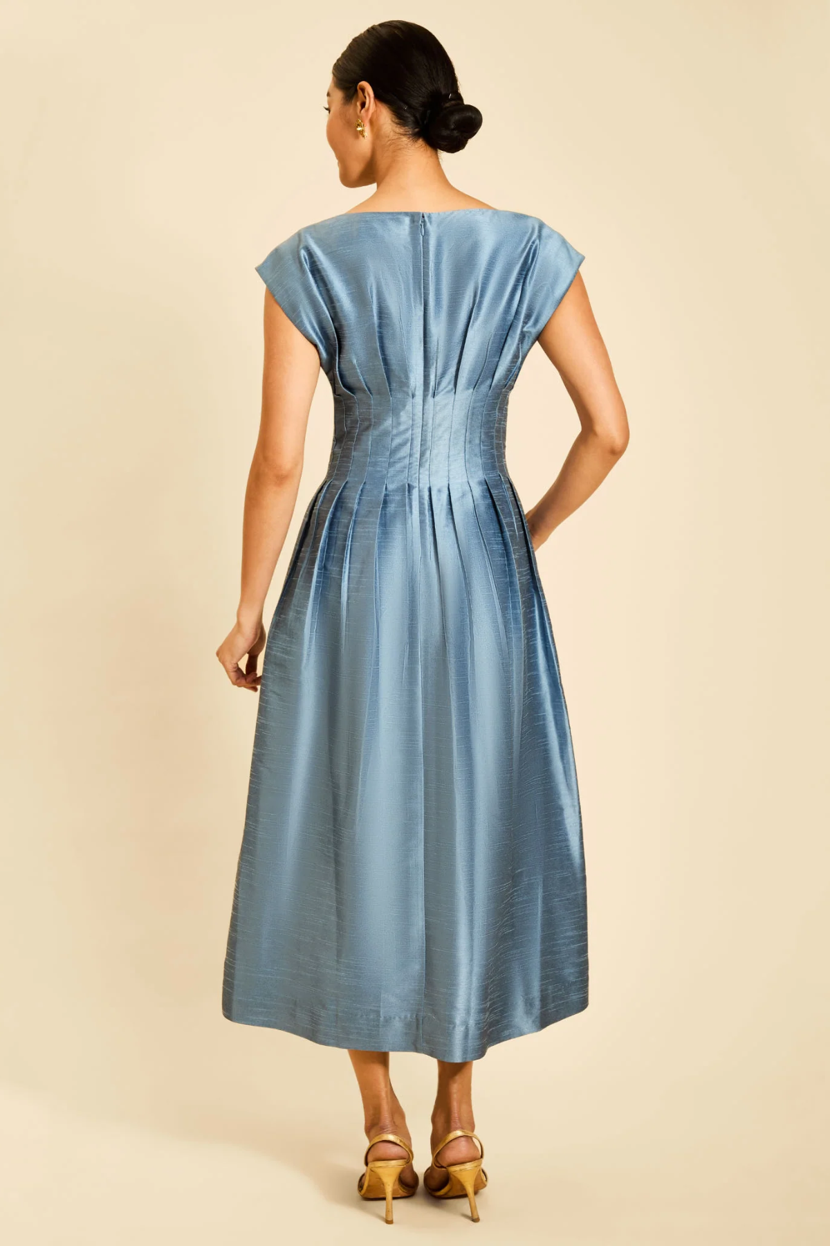 A woman shown from the back wearing a light blue midi dress with cap sleeves, a gathered waist, and a satin-like finish, paired with tan high-heeled sandals against a plain beige studio background.