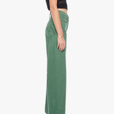 A side-profile view of a model wearing high-waisted, wide-leg sage green pants with a black sleeveless crop top and metallic high-heeled sandals, set against a plain white studio background.