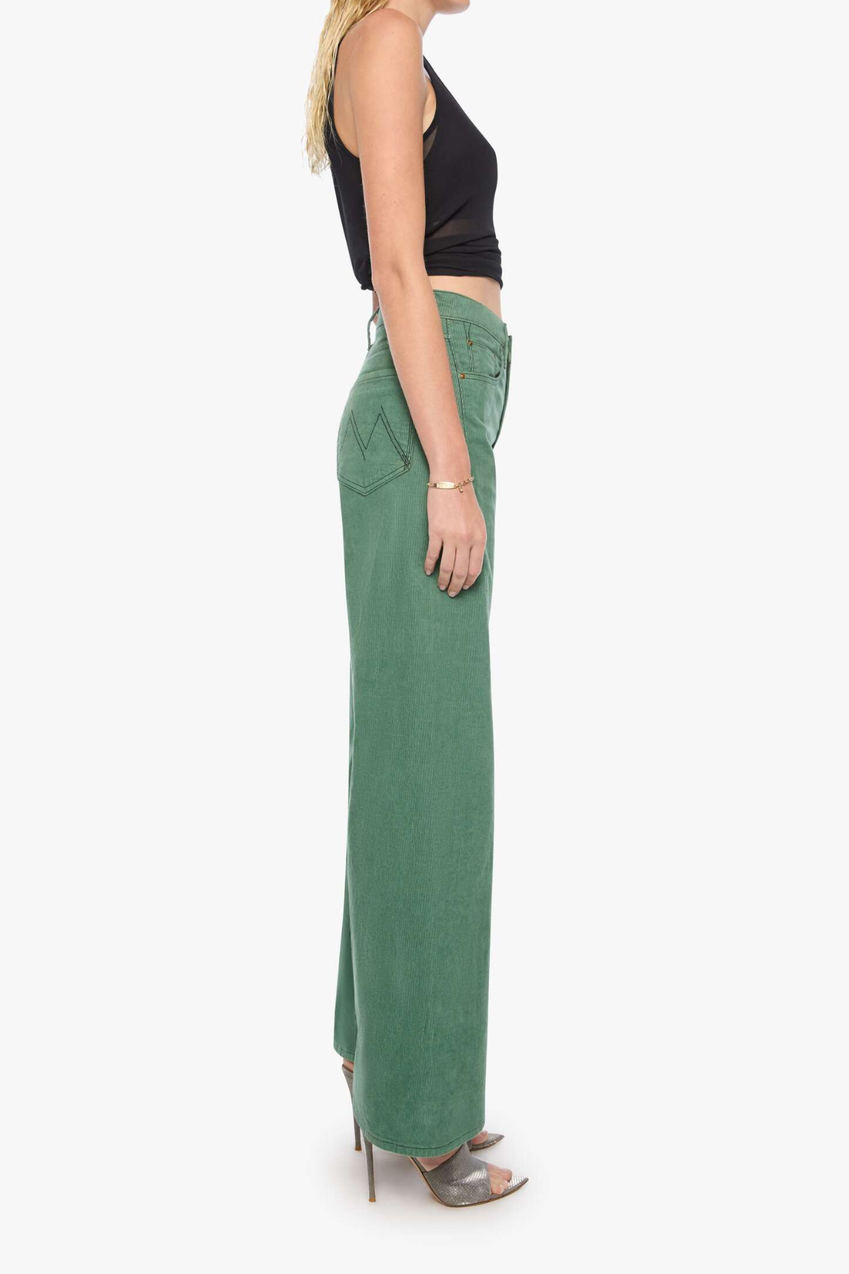 A side-profile view of a model wearing high-waisted, wide-leg sage green pants with a black sleeveless crop top and metallic high-heeled sandals, set against a plain white studio background.