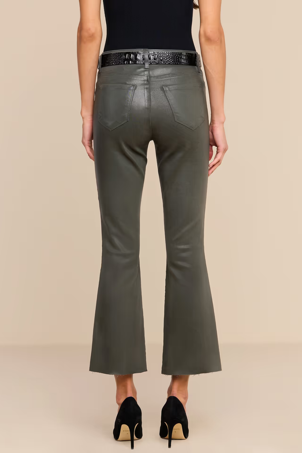 A model shown from the back wearing olive green faux-leather flare pants, a black belt, a black top, and black high heels against a solid beige background.