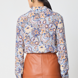 A woman shown from the back wearing a long-sleeve button-up blouse with a blue, beige, and orange floral pattern, paired with a brown faux-leather skirt, against a light gray studio background.