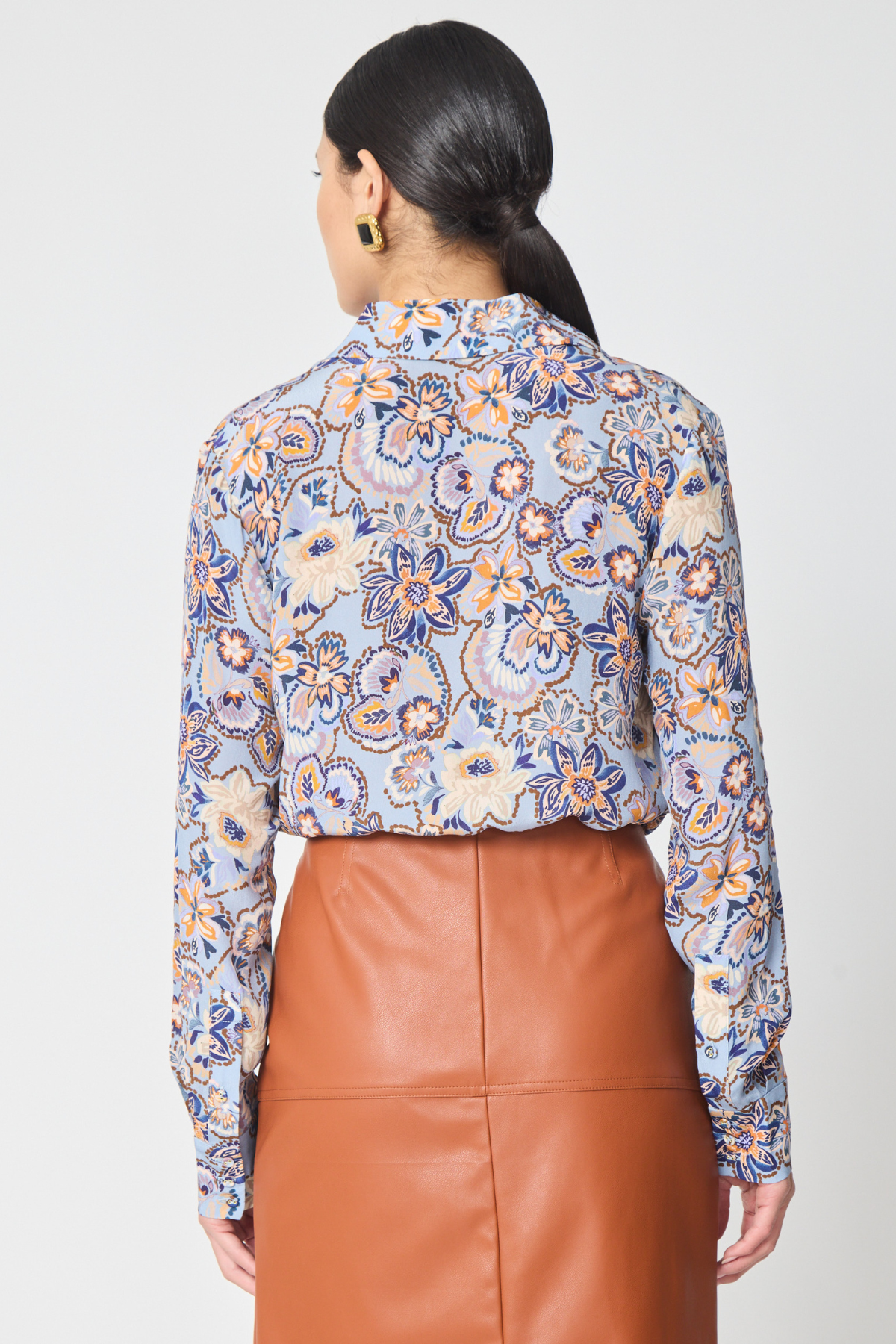 A woman shown from the back wearing a long-sleeve button-up blouse with a blue, beige, and orange floral pattern, paired with a brown faux-leather skirt, against a light gray studio background.