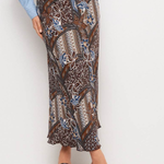 A model wearing a brown batik-patterned maxi skirt with blue and beige accents, paired with a light blue button-up shirt and black slide sandals, against a plain white studio background.