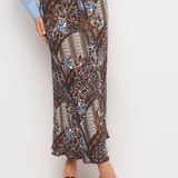 A model wearing a brown batik-patterned maxi skirt with blue and beige accents, paired with a light blue button-up shirt and black slide sandals, against a plain white studio background.