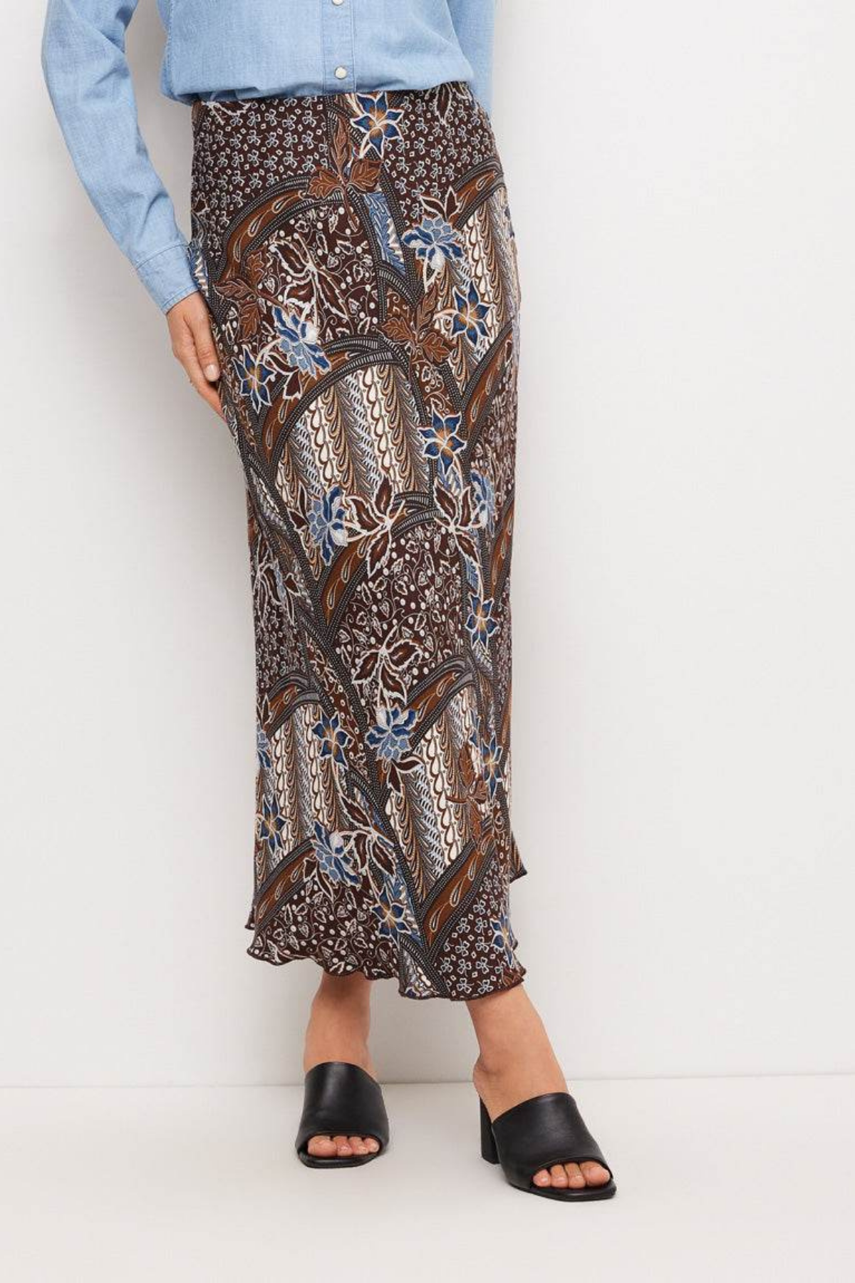 A model wearing a brown batik-patterned maxi skirt with blue and beige accents, paired with a light blue button-up shirt and black slide sandals, against a plain white studio background.