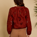 A woman viewed from behind wearing a rust-red long-sleeve blouse with black floral embroidery and tan pants, standing against a warm beige background with a plant leaf visible on the right.