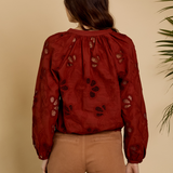 A woman viewed from behind wearing a rust-red long-sleeve blouse with black floral embroidery and tan pants, standing against a warm beige background with a plant leaf visible on the right.