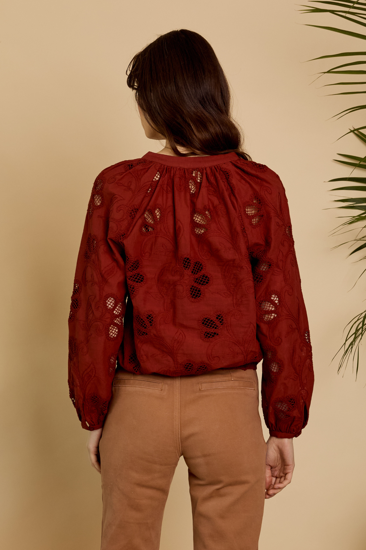 A woman viewed from behind wearing a rust-red long-sleeve blouse with black floral embroidery and tan pants, standing against a warm beige background with a plant leaf visible on the right.