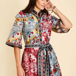 A woman modeling a short, button-front dress with a colorful paisley and floral print, featuring a tie waist belt and elbow-length sleeves, against a solid beige background.