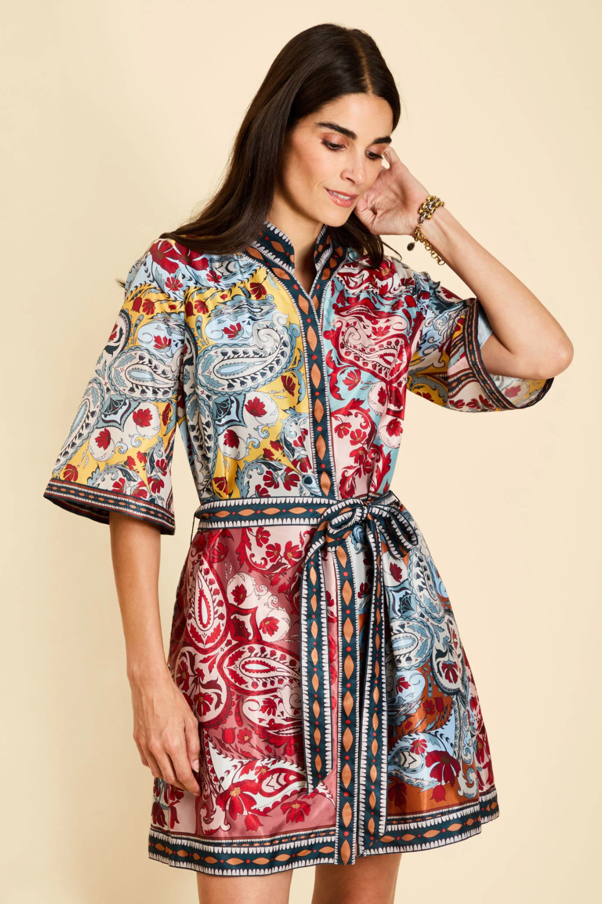 A woman modeling a short, button-front dress with a colorful paisley and floral print, featuring a tie waist belt and elbow-length sleeves, against a solid beige background.