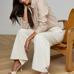 Woman sitting on a wooden chair wearing a beige shirt and white pants.