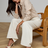 Woman sitting on a wooden chair wearing a beige shirt and white pants.