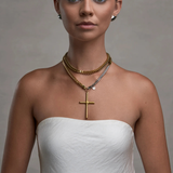 Woman wearing a gold cross necklace and multiple layered necklaces against a neutral background