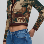 A cropped long-sleeve button-front top with a scenic landscape print in beige, brown, and dark green, worn by a model with blue jeans against a light gray studio background.