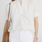 Woman wearing a white blouse and white pants, holding a woven bag against a plain background