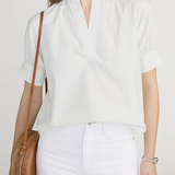 Woman wearing a white blouse and white pants, holding a woven bag against a plain background