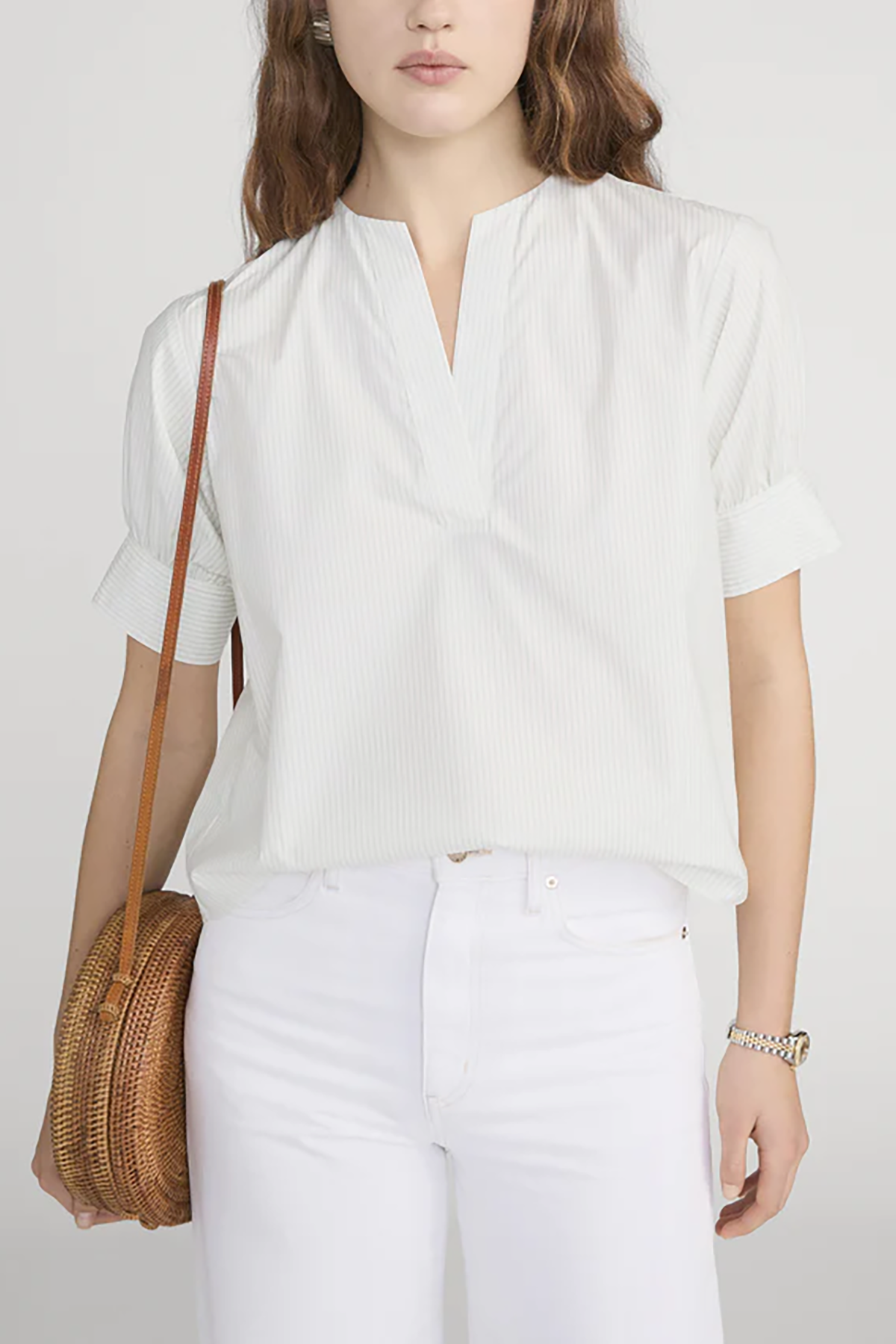 Woman wearing a white blouse and white pants, holding a woven bag against a plain background