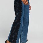 A model wears high-waisted wide-leg blue denim jeans featuring a prominent black floral lace panel down the left leg, styled with black pointed-toe stiletto heels against a light gray studio background.