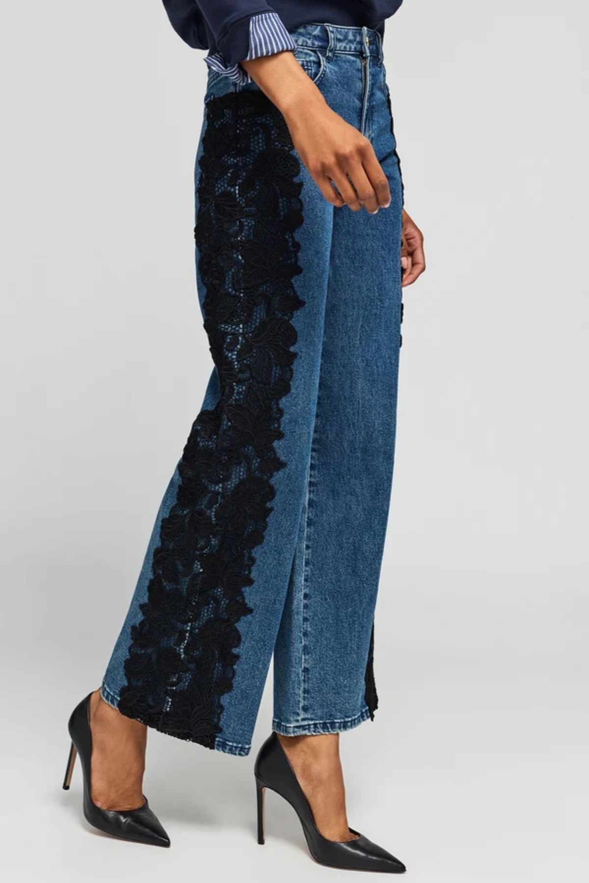 A model wears high-waisted wide-leg blue denim jeans featuring a prominent black floral lace panel down the left leg, styled with black pointed-toe stiletto heels against a light gray studio background.