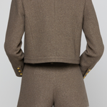 A model shown from the back wearing a matching cropped blazer and shorts set in a textured taupe-brown fabric, featuring gold button accents on the cuffs.
