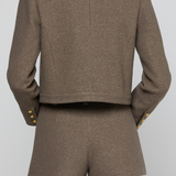 A model shown from the back wearing a matching cropped blazer and shorts set in a textured taupe-brown fabric, featuring gold button accents on the cuffs.