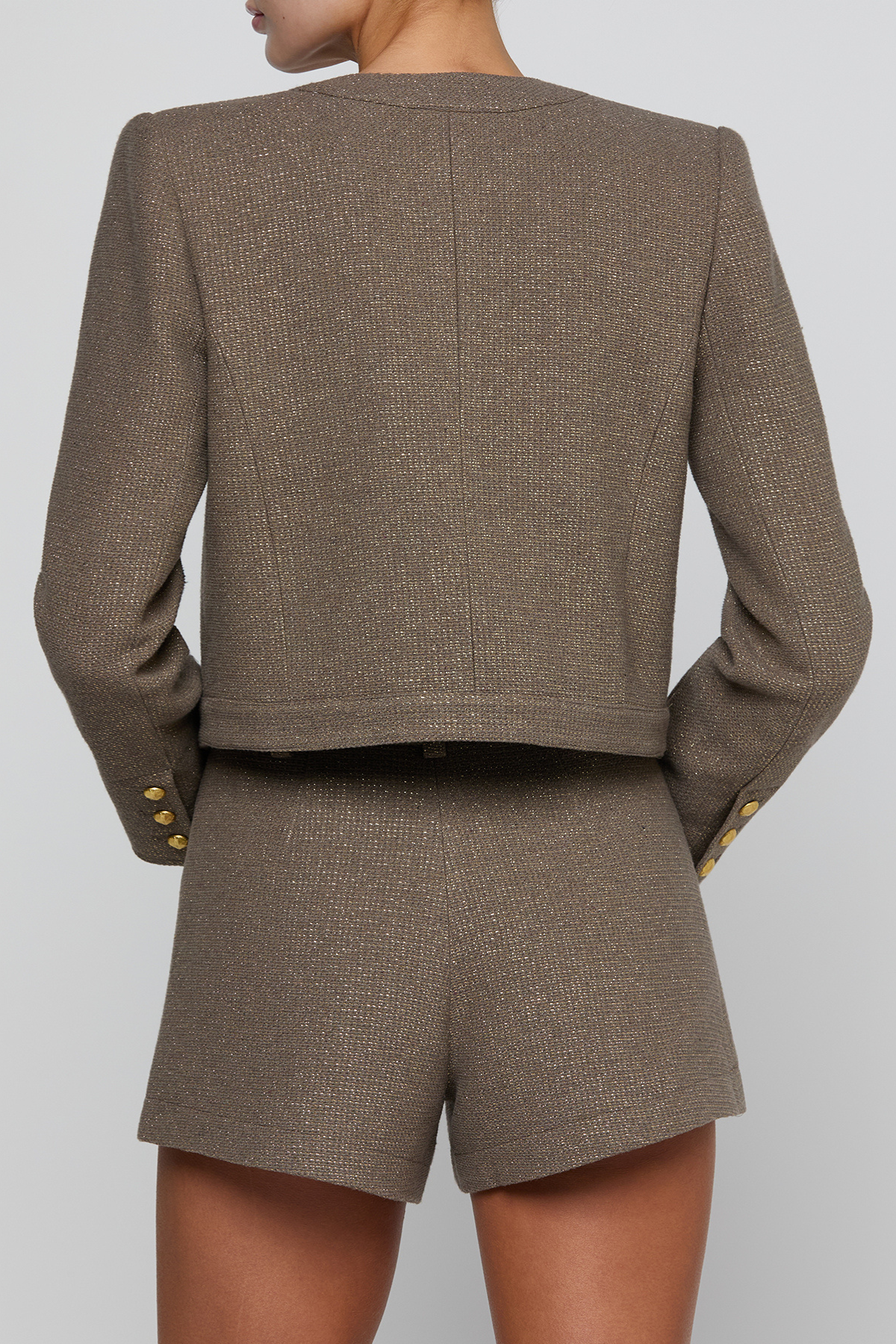 A model shown from the back wearing a matching cropped blazer and shorts set in a textured taupe-brown fabric, featuring gold button accents on the cuffs.