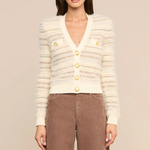 A woman modeling a cream-colored textured knit cardigan with gold buttons, paired with brown corduroy pants, against a solid beige studio background.