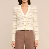 A woman modeling a cream-colored textured knit cardigan with gold buttons, paired with brown corduroy pants, against a solid beige studio background.