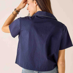 A woman shown from the back wearing a navy blue short-sleeve collared shirt and light blue jeans against a plain light beige background.