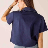 A woman shown from the back wearing a navy blue short-sleeve collared shirt and light blue jeans against a plain light beige background.
