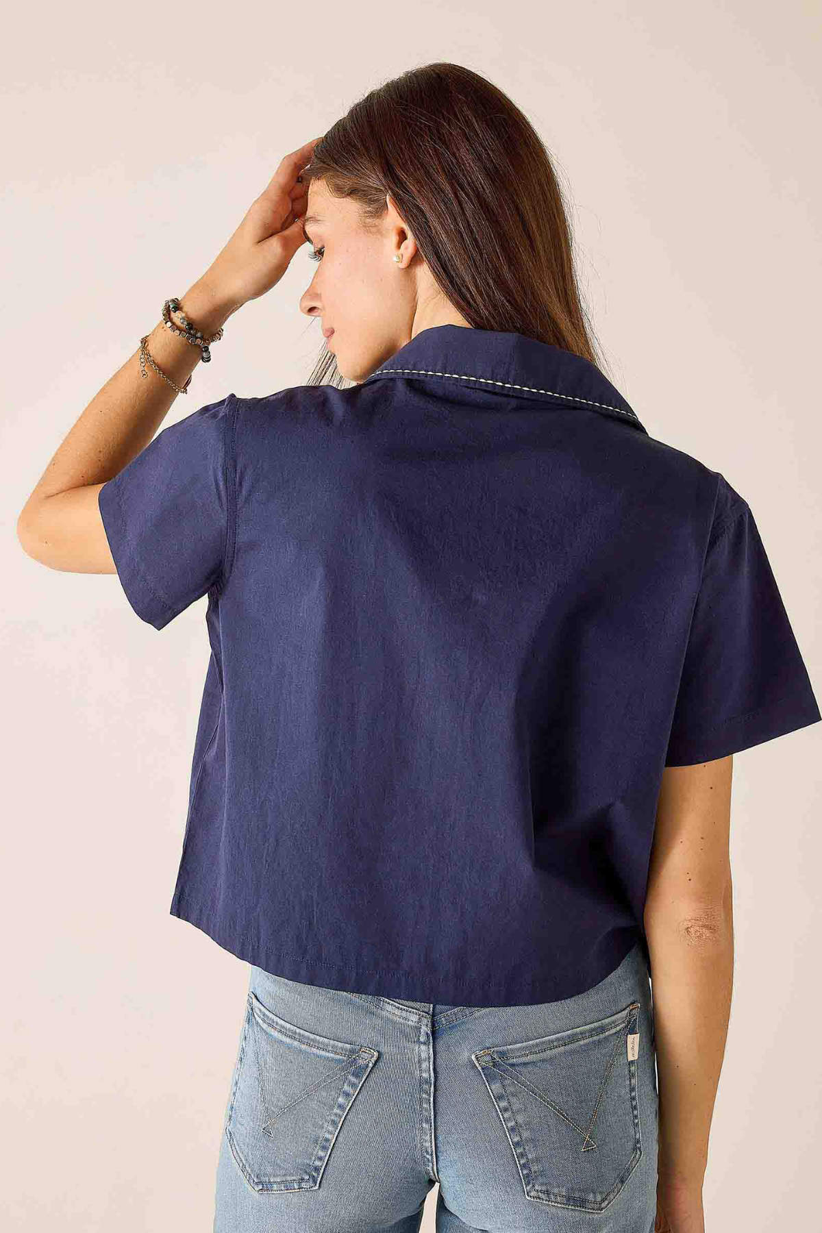 A woman shown from the back wearing a navy blue short-sleeve collared shirt and light blue jeans against a plain light beige background.
