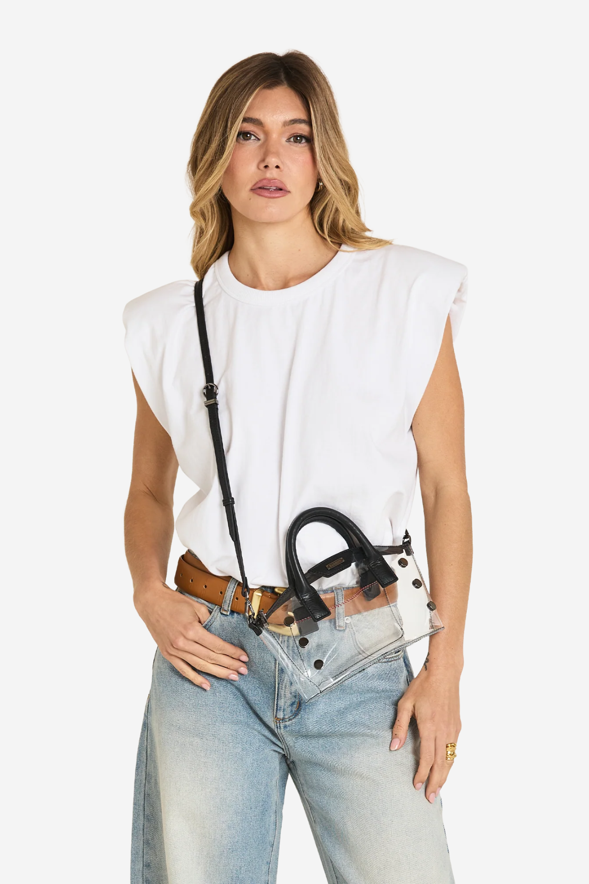A woman modeling a white sleeveless top and light-wash jeans, wearing a black studded shoulder bag with a long strap and a brown belt.