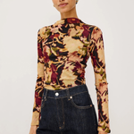 A woman modeling a fitted long-sleeve mock-neck top with a multicolor abstract floral pattern in beige, burgundy, olive green, and black, paired with dark blue jeans against a plain light gray studio background.