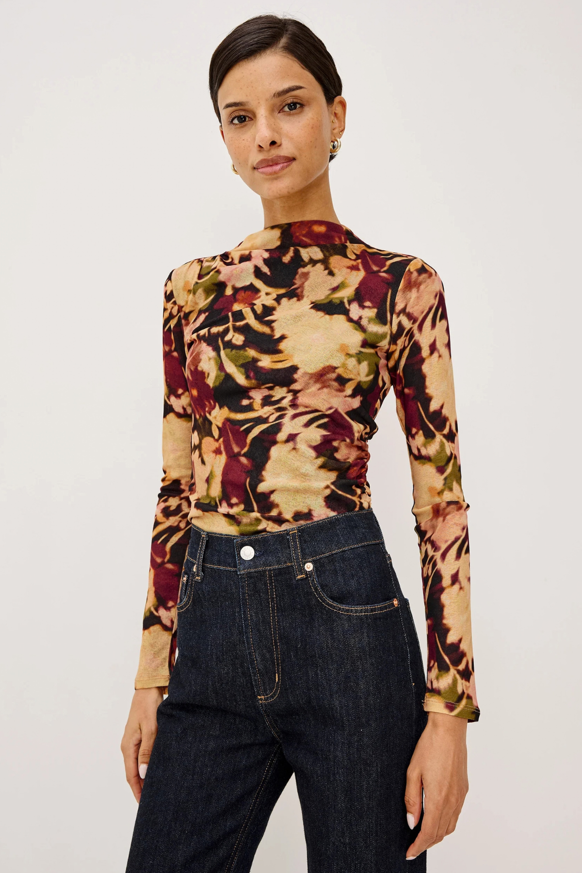 A woman modeling a fitted long-sleeve mock-neck top with a multicolor abstract floral pattern in beige, burgundy, olive green, and black, paired with dark blue jeans against a plain light gray studio background.