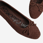 A pair of dark brown woven ballet flats with bow details, shown from a top-down angle on a light gray background. The brand name 'Saskatoon' is visible on the insole.
