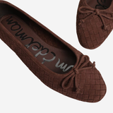 A pair of dark brown woven ballet flats with bow details, shown from a top-down angle on a light gray background. The brand name 'Saskatoon' is visible on the insole.