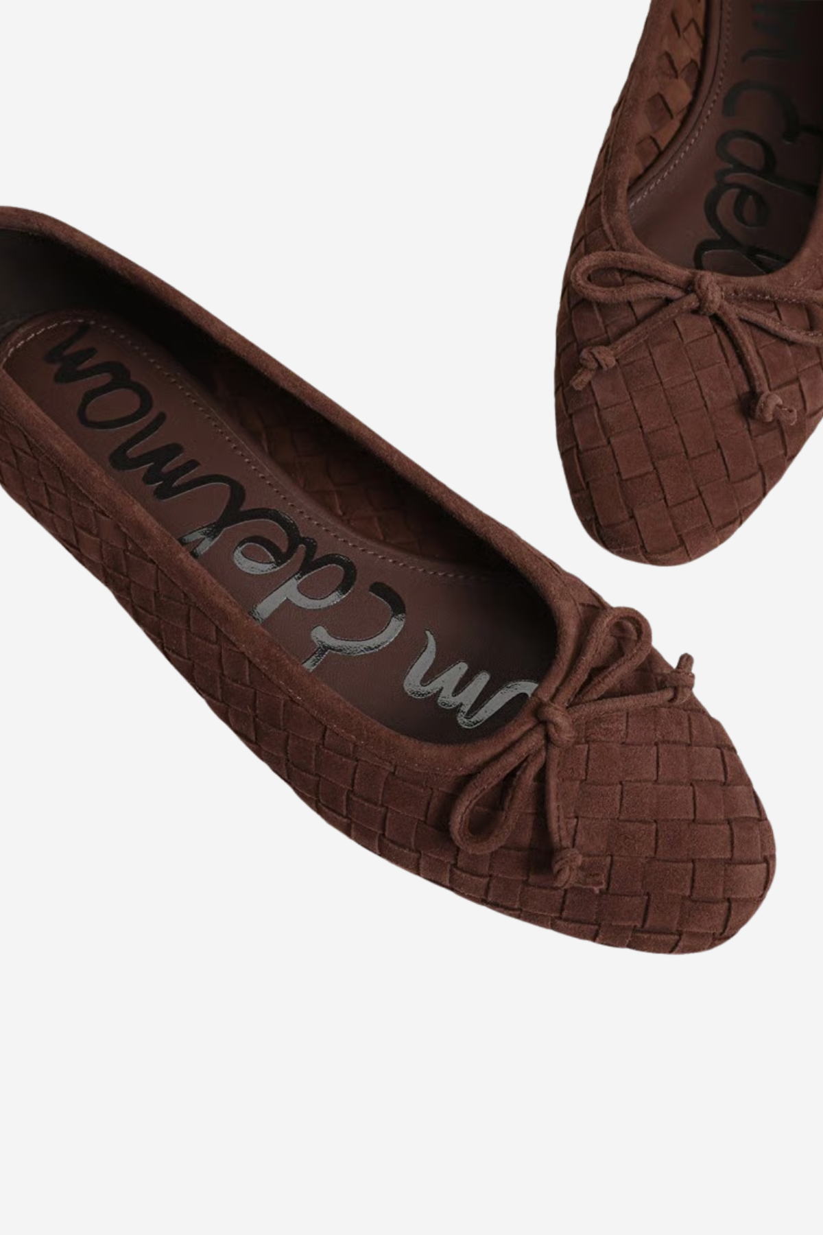 A pair of dark brown woven ballet flats with bow details, shown from a top-down angle on a light gray background. The brand name 'Saskatoon' is visible on the insole.