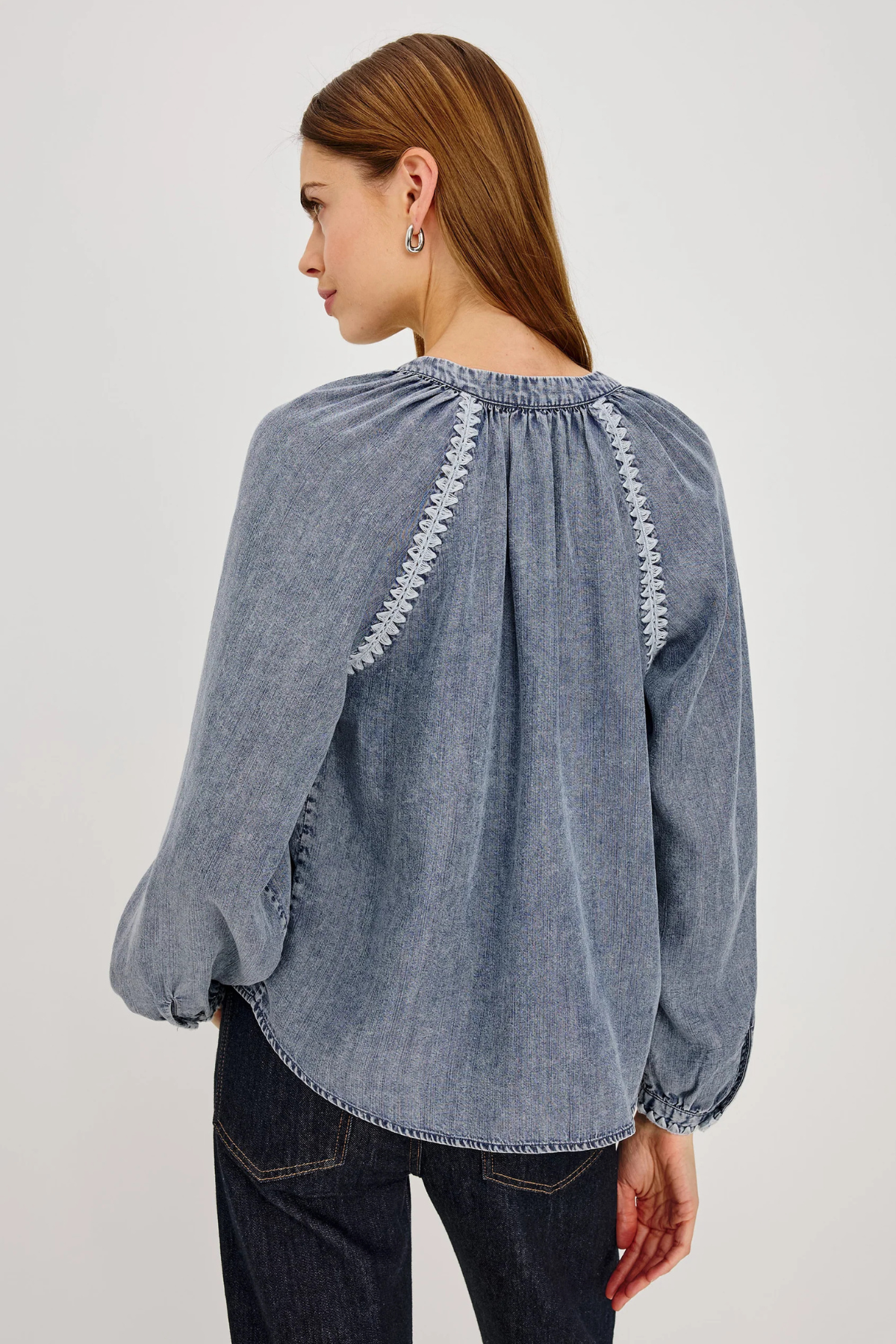 A woman shown from the back wearing a dusty blue denim long-sleeve blouse with white embroidered details on the shoulders, paired with dark blue jeans, against a light gray studio background.