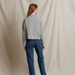 A woman stands with her back to the camera wearing a light gray cropped sweater and blue jeans, standing against a draped beige fabric backdrop.
