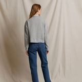 A woman stands with her back to the camera wearing a light gray cropped sweater and blue jeans, standing against a draped beige fabric backdrop.
